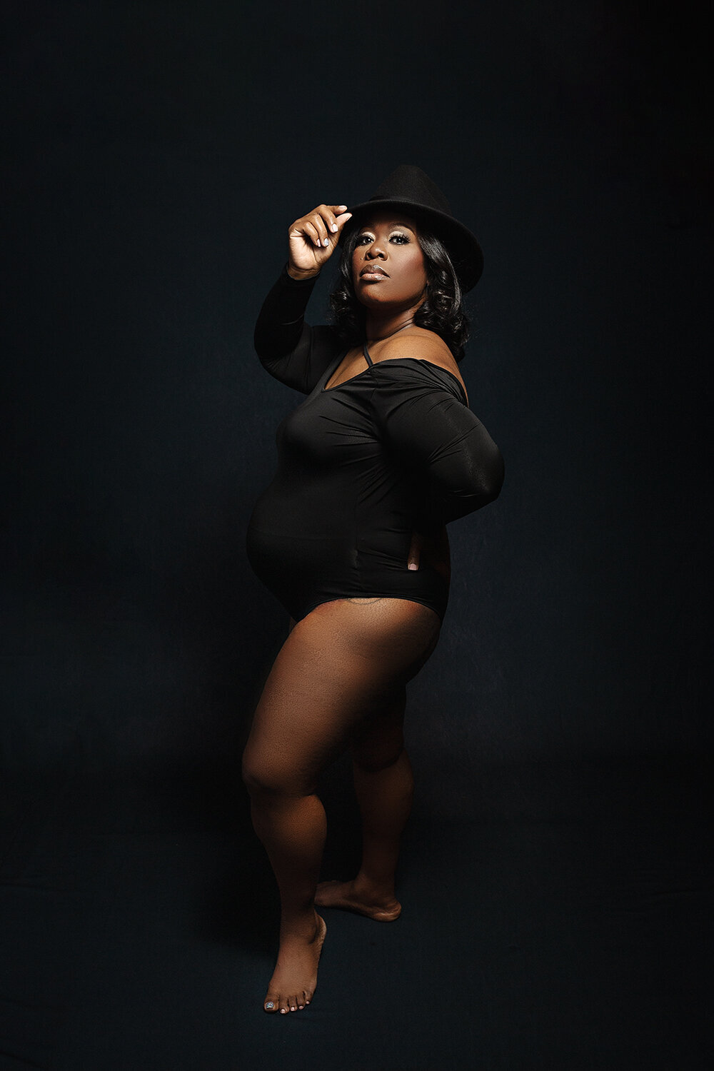 A mother to be in a black long-sleeve one-piece maternity suit tips her hat with a leg popped