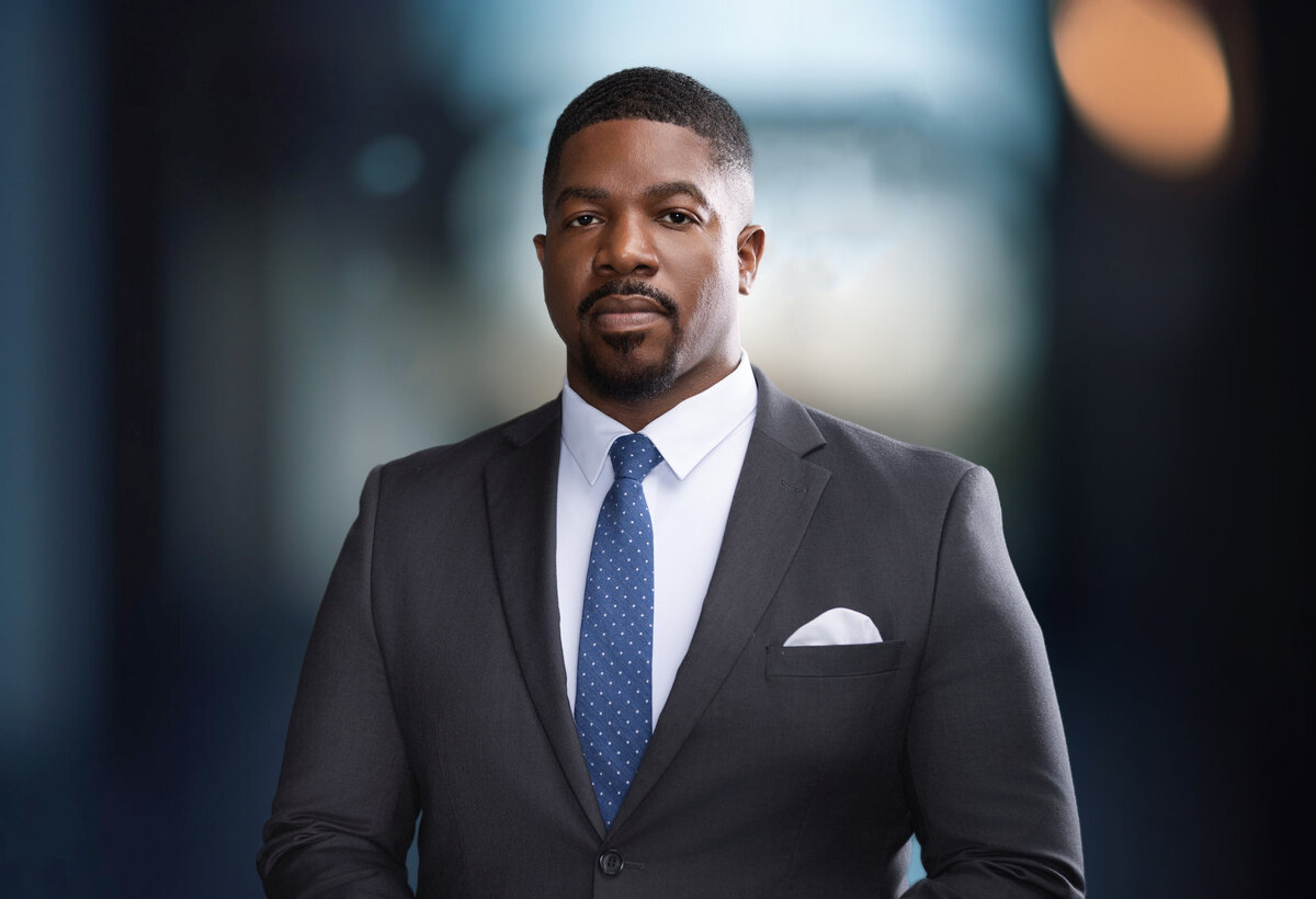A professional headshot of Omar Waller, showcasing confidence and professionalism. He is wearing a dark gray suit, a crisp white shirt, and a blue patterned tie, complemented by a white pocket square. The blurred modern background adds depth, emphasizing his poised demeanor. Captured in Cincinnati, this high-quality portrait is ideal for professional branding, highlighting the expertise of a skilled portrait photographer and studio.