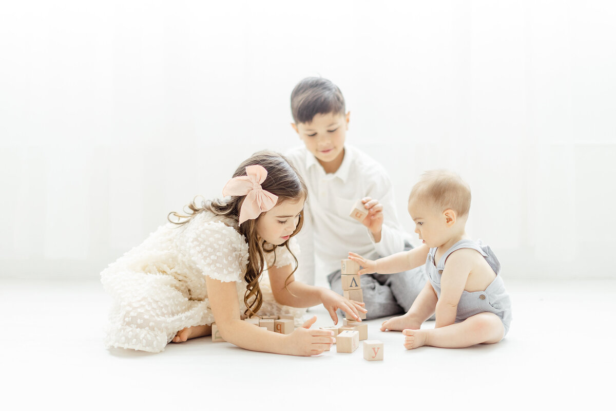 3 young siblings playing on the floor of a photography studio with wooden blocks.