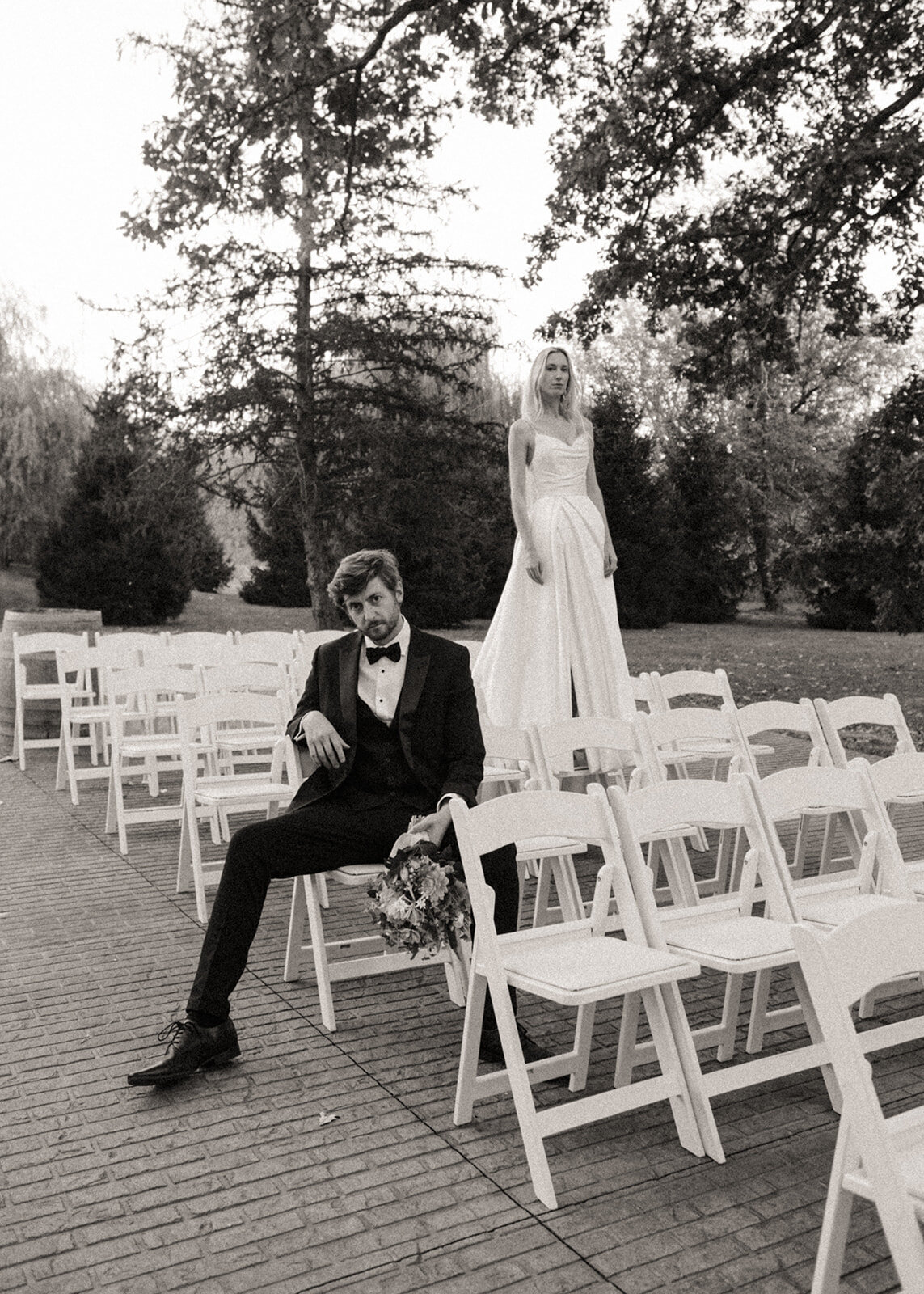 A bride standing and a groom sitting on white chairs outdoors at Park Farm Winery, both dressed in wedding attire, in a black and white photo.