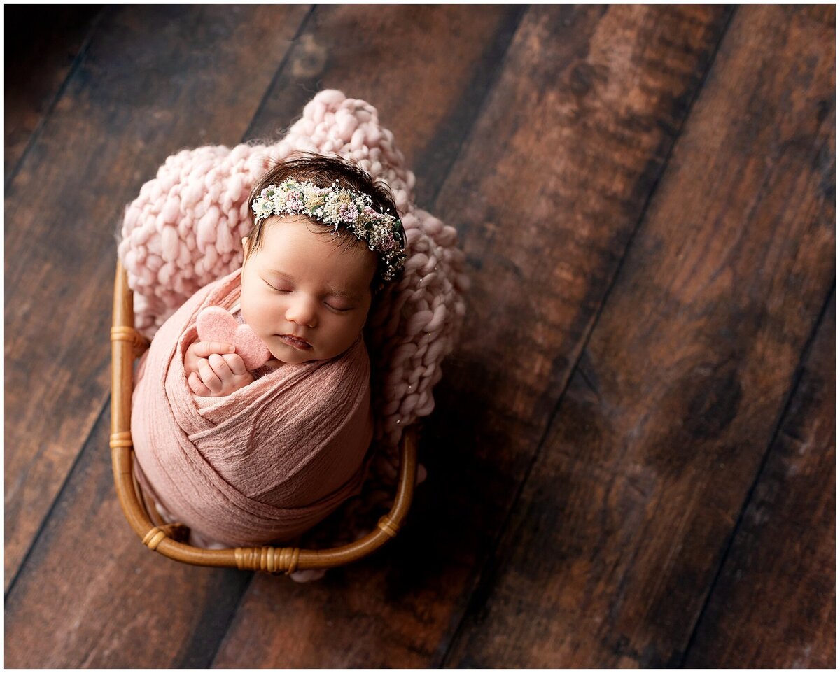 A newborn baby girl wrapped in pink swaddle holding a heart and flower headband on her head. The baby is laying a basket.