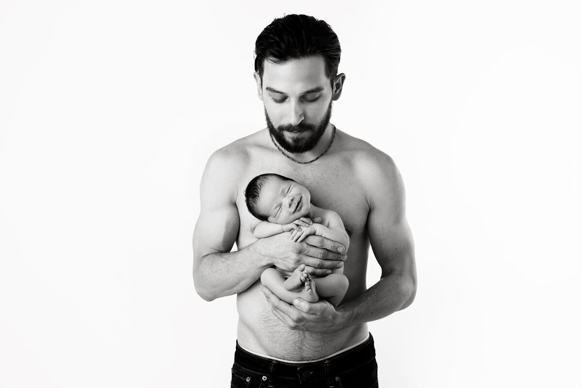 A shirtless new father stands in a studio cradling his newborn baby against his chest