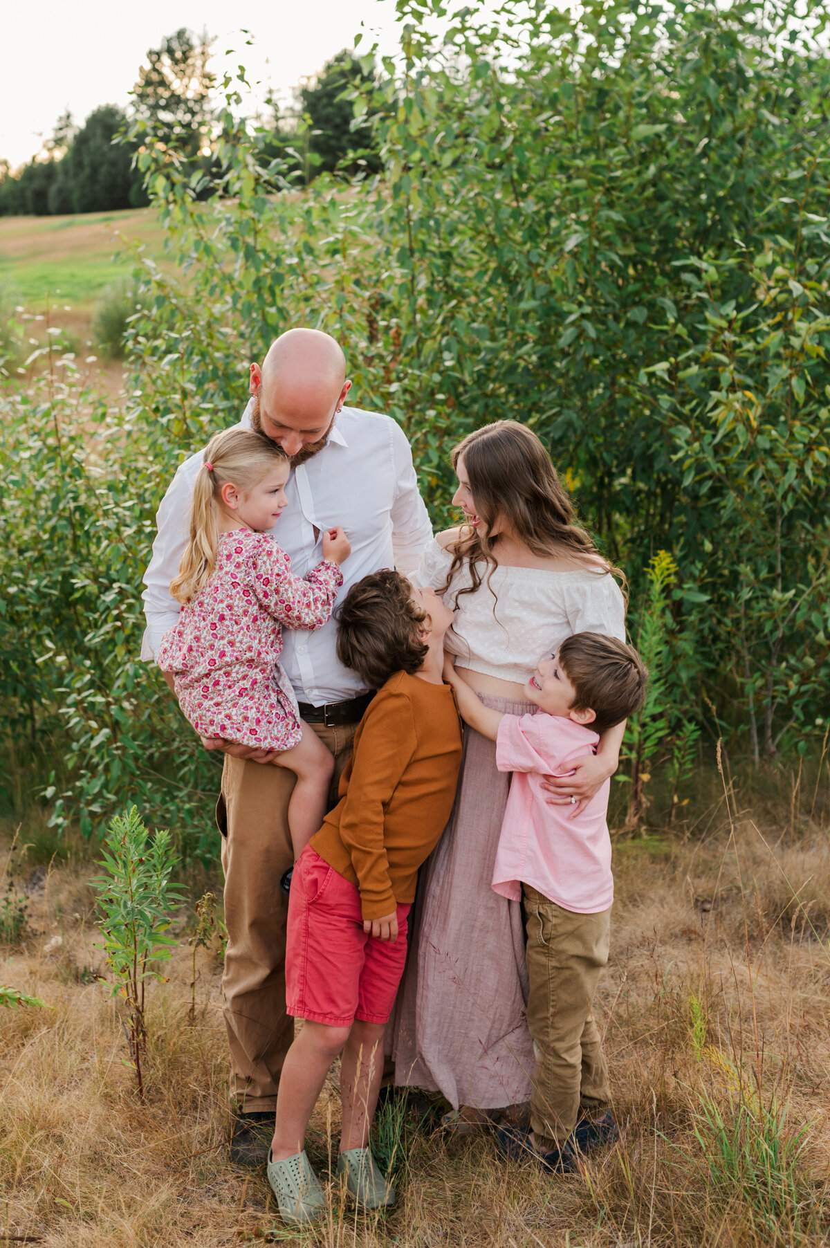 Family close together dressed nice, cuddled in looking at each other and smiling. Family photos are in a greenery field  in the back round.
