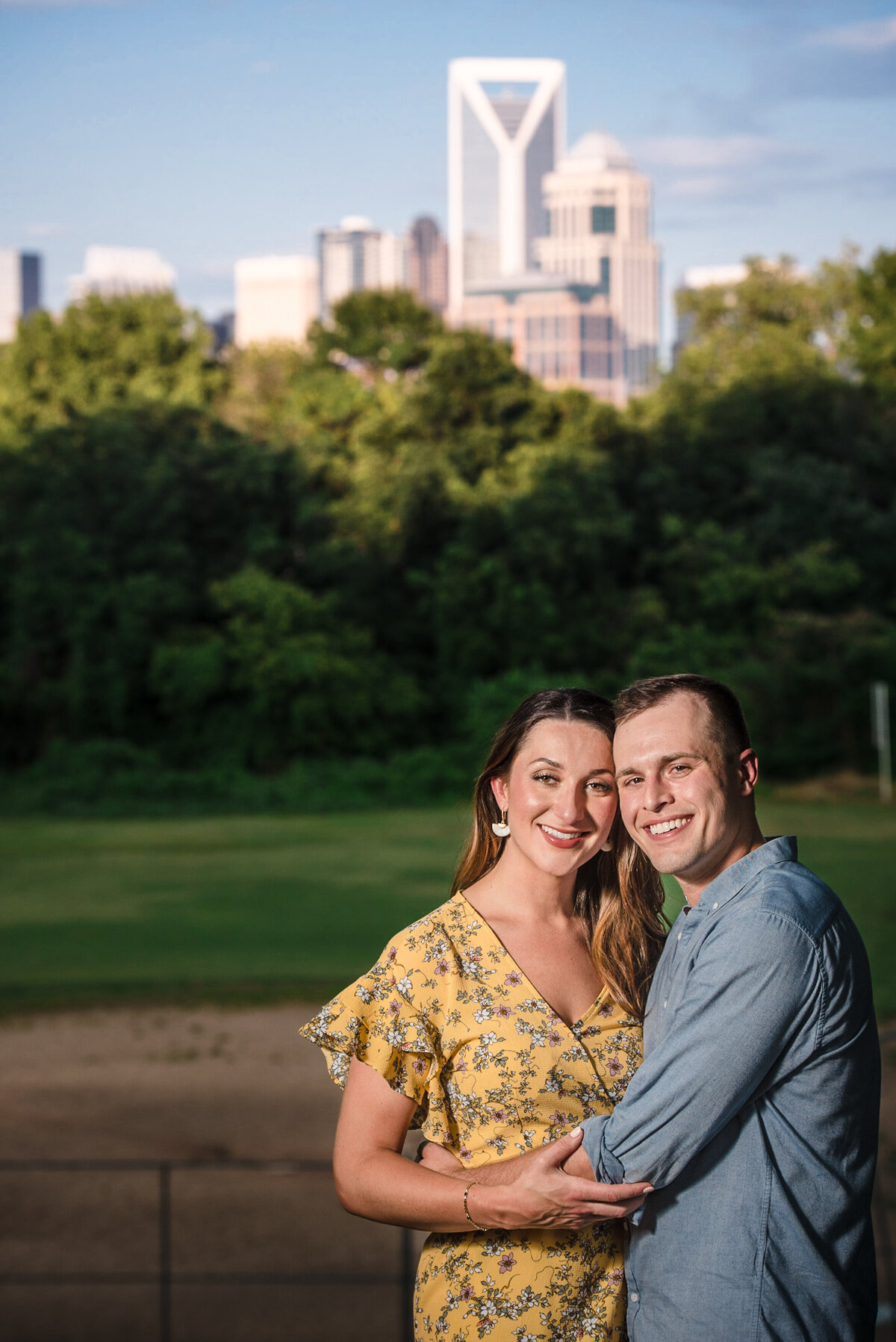 Engaged-couple-smiling-and-looking-at-the-camera-with-the-Charlotte-skyline-in-the-background-at-Bryant-Park