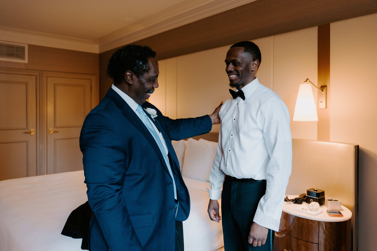 Two men in formal attire smile warmly in a hotel room. One rests his hand on the other's shoulder, conveying a sense of support and celebration.