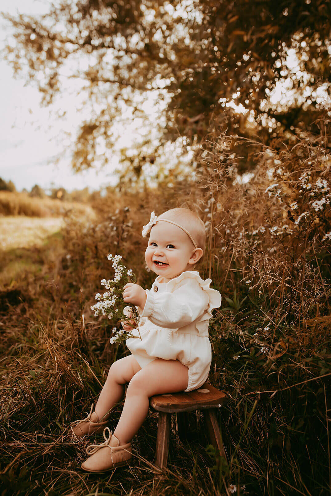 a one year old girl sitting on a stool holding wild flowers