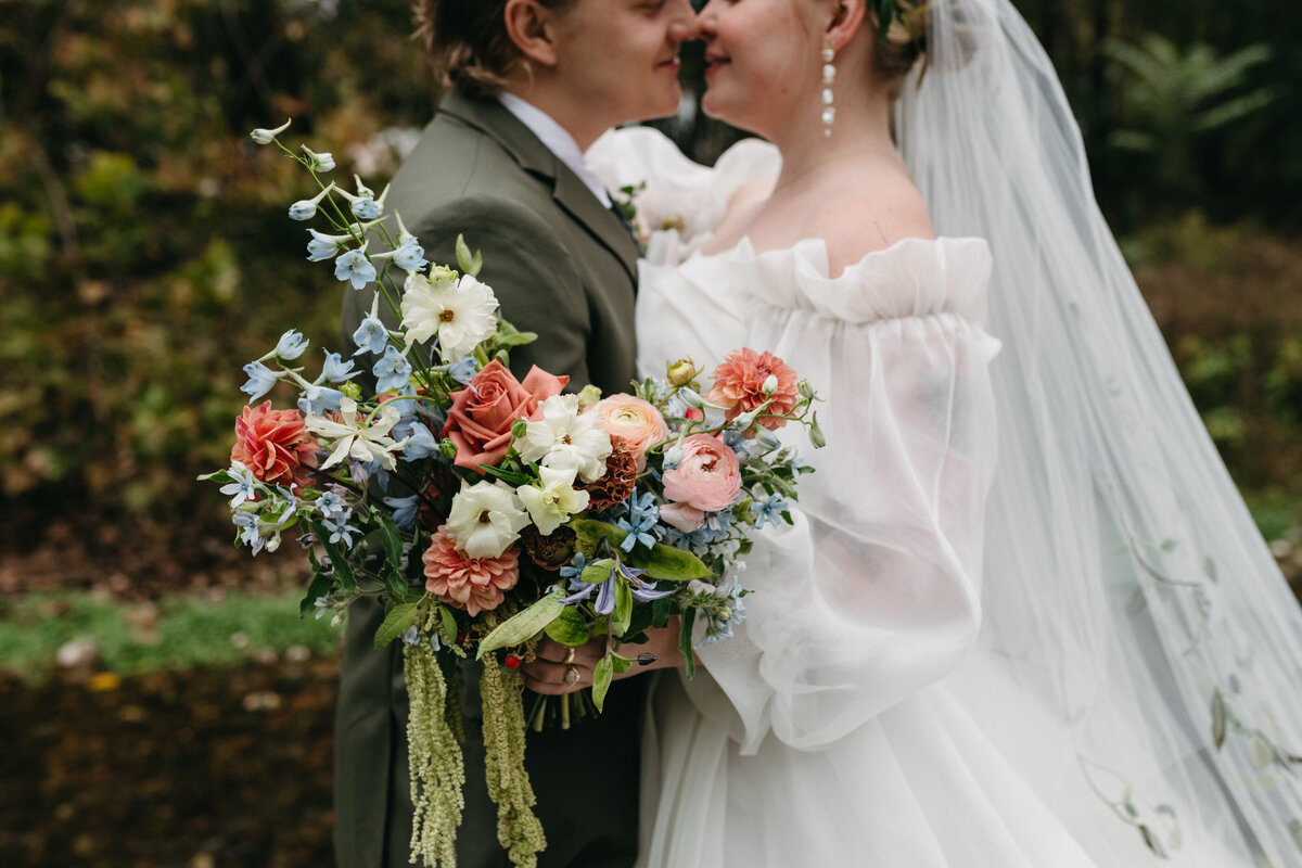 Close-up of the bride’s garden-inspired bouquet featuring soft pink ranunculus, rust roses, blue delphinium, creamy lisianthus, and trailing amaranthus, arranged in a whimsical, organic style by a Northwest Arkansas wedding florist.