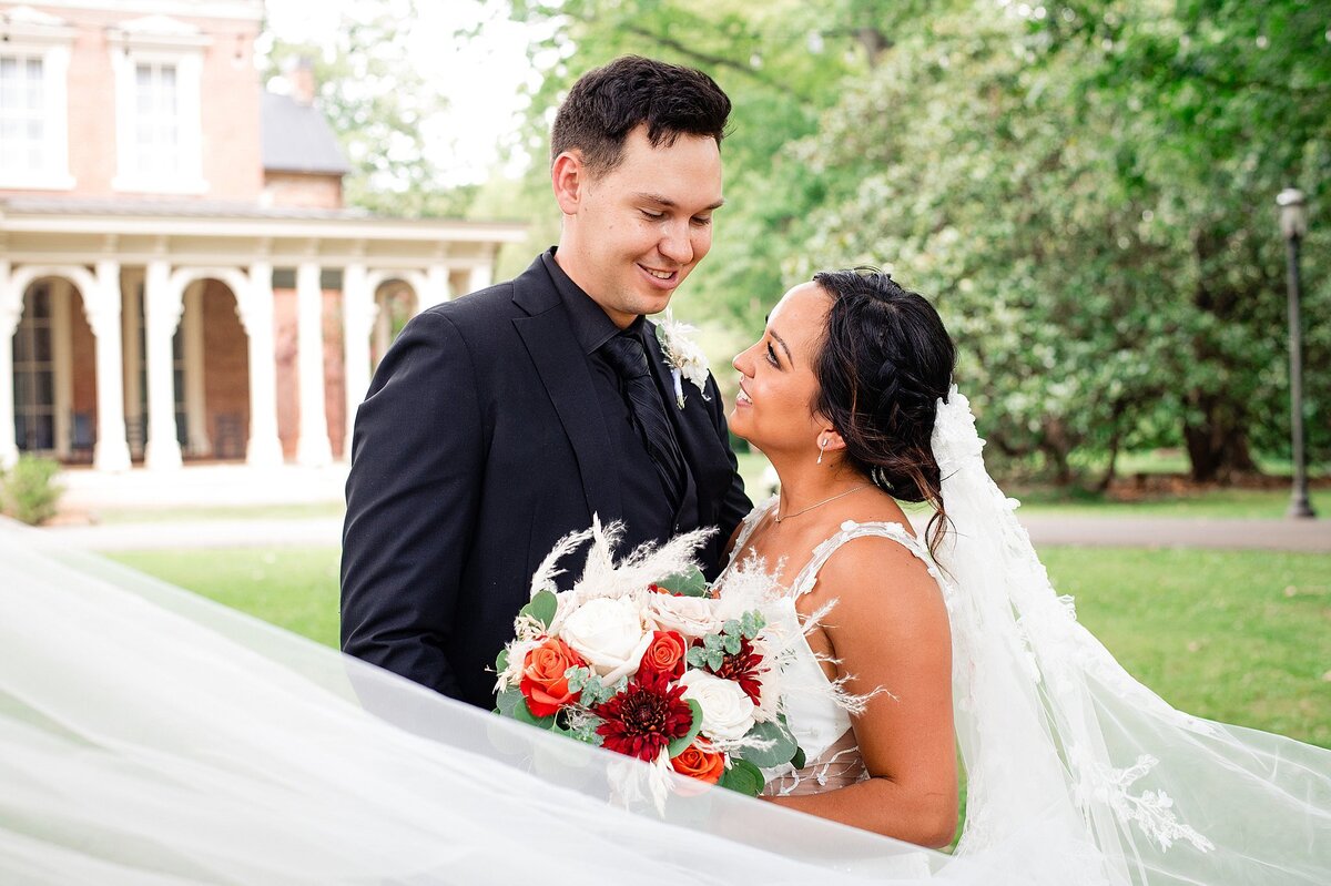 Standing in front of Oaklands Mansion. the bride's long veil flows in the wind as she looks up at the groom who is wearing a black suit with a black shirt. The bride is holding a boho bouquet with orange roses, crimson dahlia, ivory roses, silver dollar eucalyptus and pampas grass.