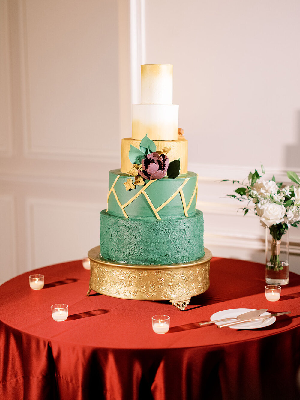 Stunning five-tier wedding cake in emerald green, white, and gold, displayed on a gold cake stand with a red linen tablecloth for a December wedding at the Don Cesar Hotel, St. Pete Beach, FL.