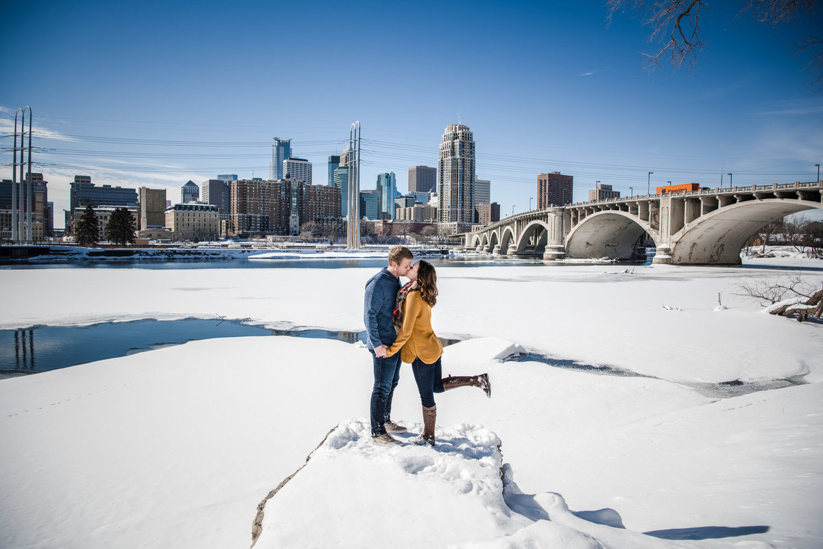 Man and woman kiss on a rock covered in snow in front of a frozen river.
