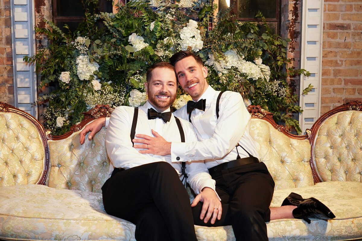 Two grooms in classic attire sit on antique couch with extravagant greenery and white flowers in the background on wedding day.