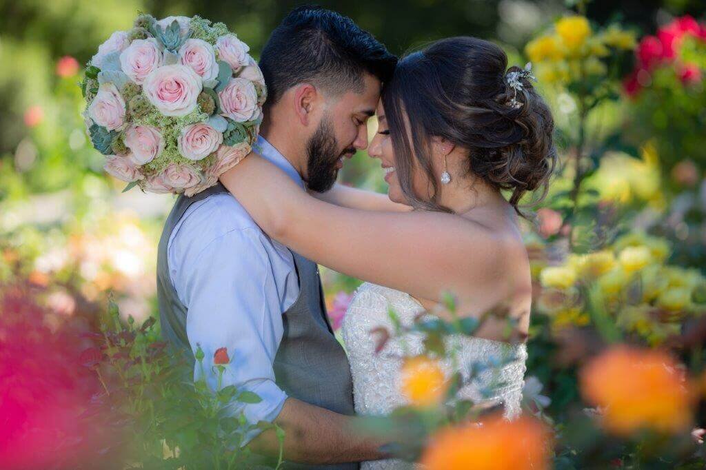 Bride and groom touch heads in the midst of pink and orange flowers smiling at the Capitol rose Gardens in Sacramento.