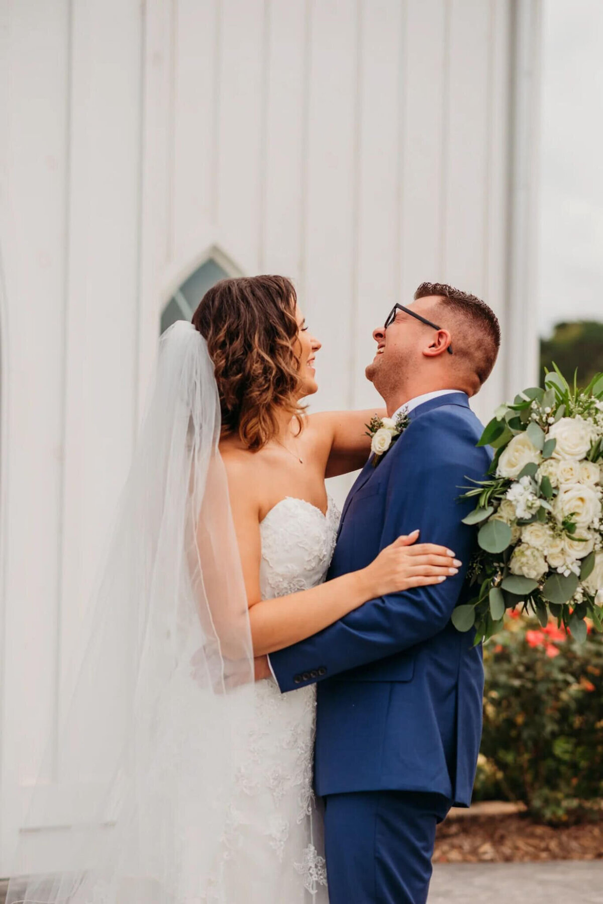 photo of a bride and groom hugging and laughing while standing in front of a white building