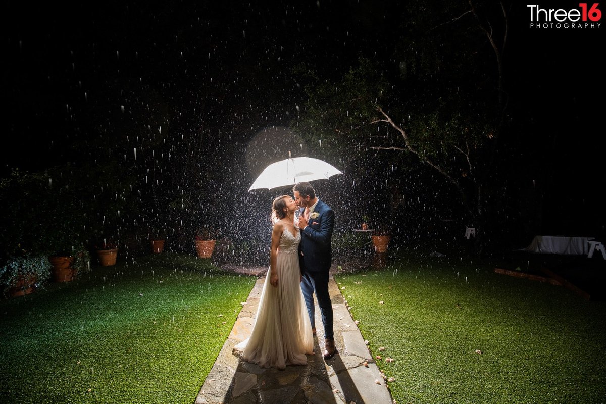 With a spotlight on them Bride and Groom share a kiss at night under an umbrella as the rain comes down