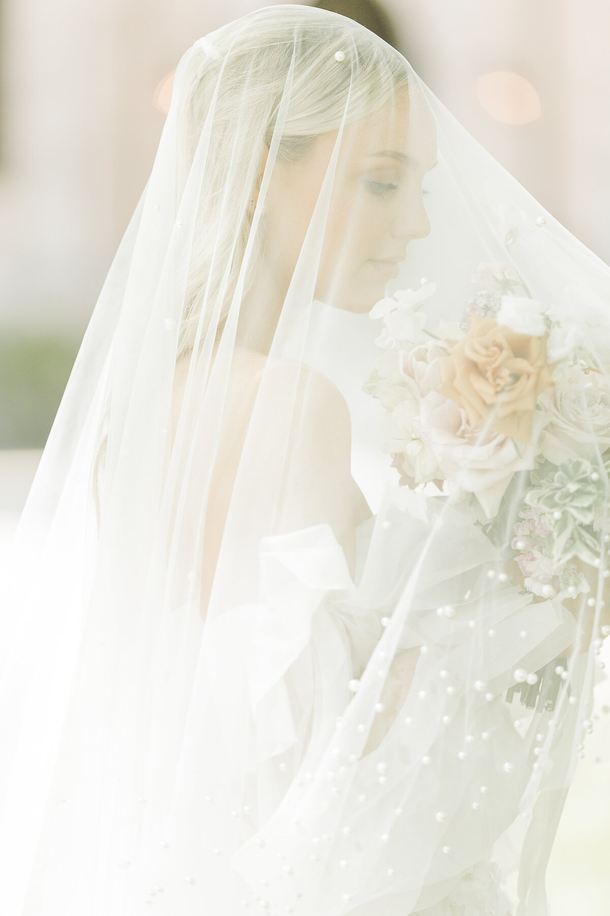 A bride stands with her veil pulled over her head. Her bouquet is under her veil. She is looking down at her flowers. Captured at golden hour for a soft, airy image. Captured by best North Shore Boston Wedding Photographer Lia Rose Weddings.