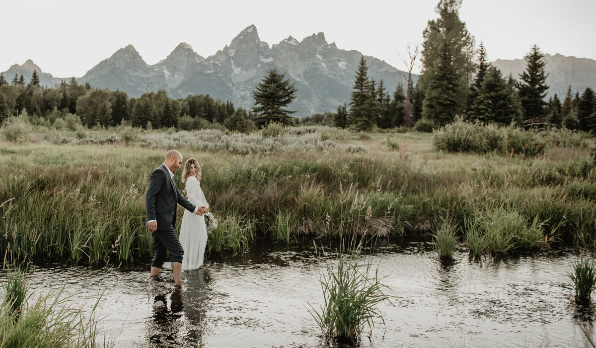 wyoming elopement in the Tetons with the bride who is wearing a classic white gown and her groom are walking in a creek in the fields in front of the Tetons