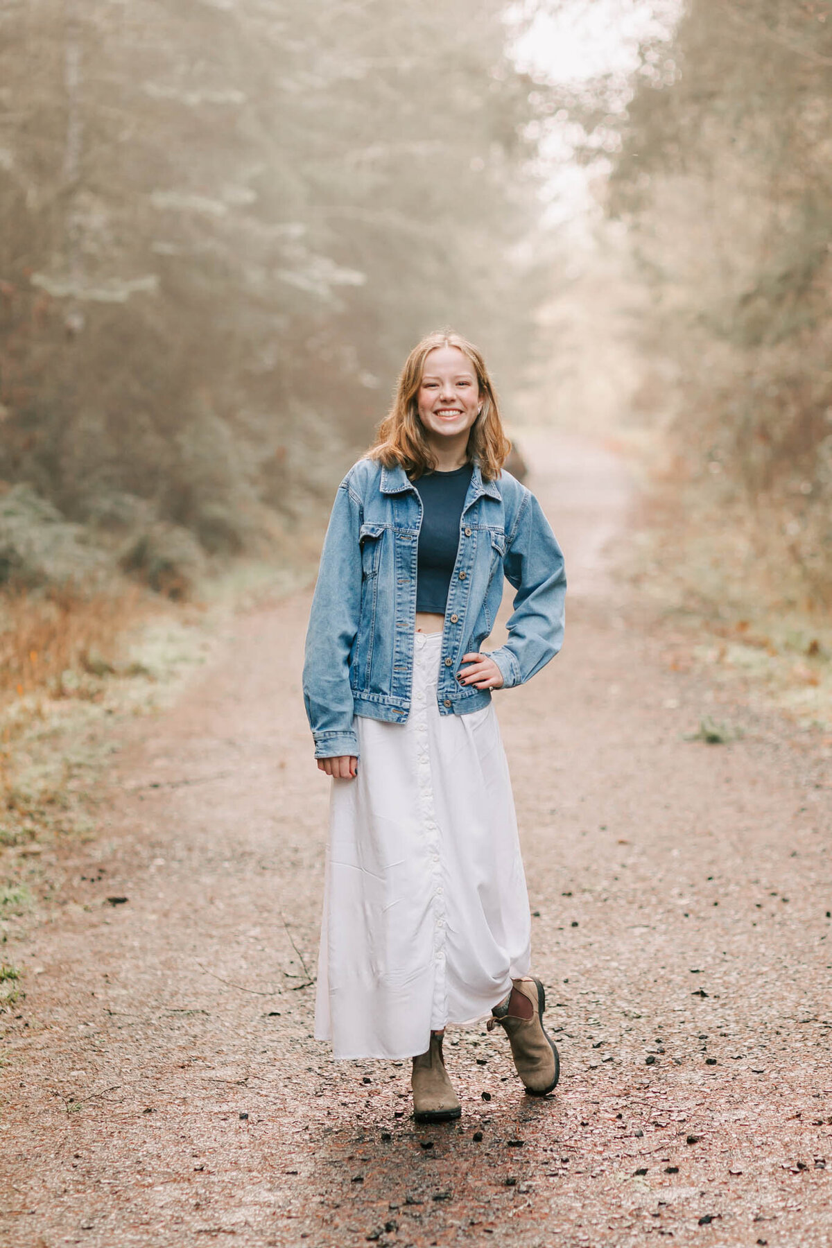 A young woman poses in the path at Fitton Green in Corvallis, Oregon for senior portraits with Jen White Photography. She's standgin with her anklyes crossed and one hand on her hip in a white skirt, tan boots, blue shirt, and jean jacket.