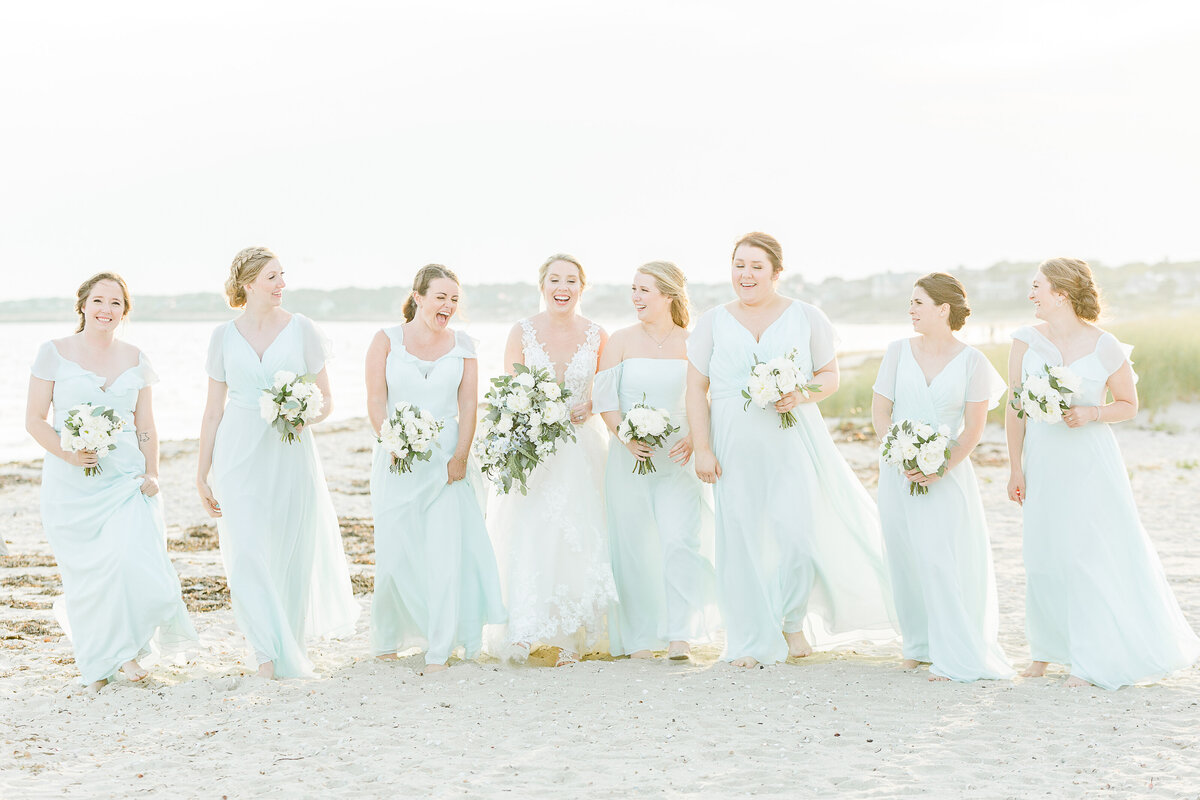 A bride and her seven bridesmaids are walking along the white sandy beaches of the Wychmere Beach Club in Harwich Port, MA for a casual bridal photo. The bride and bridesmaids are all looking and laughing at each other for this joyful photo. Captured by MA wedding photographer Lia Rose Weddings