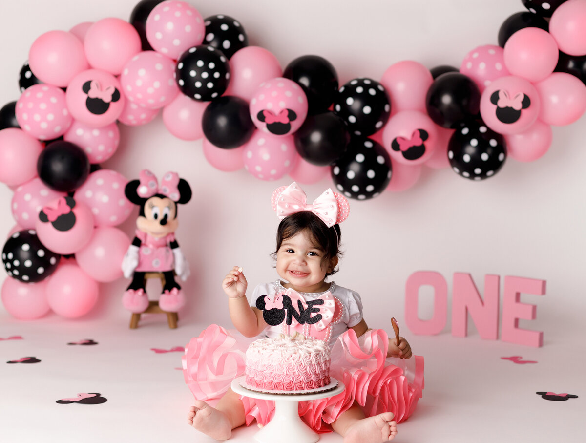 "Minnie Mouse" theme cake smash in West Palm Beach and Boca Raton photography studio. Baby girl is wearing pink glittery mouse ears and pink tutu, smiling at the camera. In the background is a pink and black balloon arch with polka dot and minnie mouse balloons. Baby is about to touch the cake for the first time.