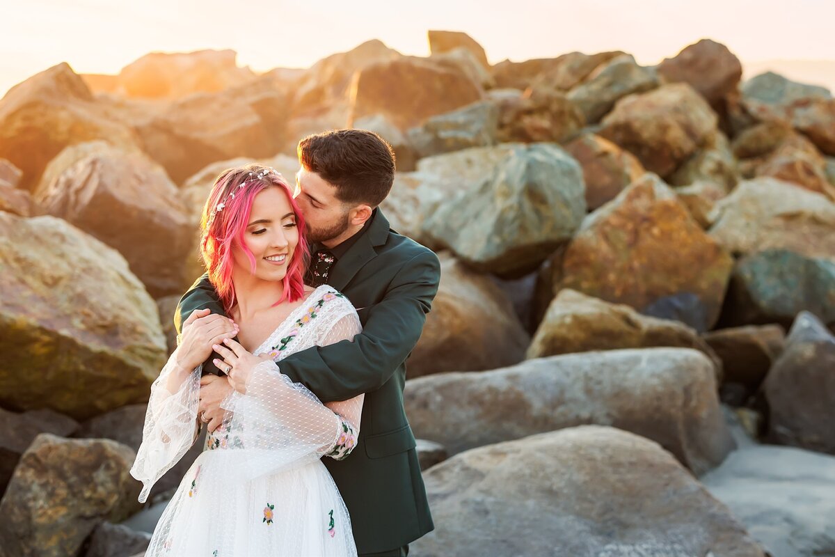 Groom hugging bride in front of rock jetty in Coronado