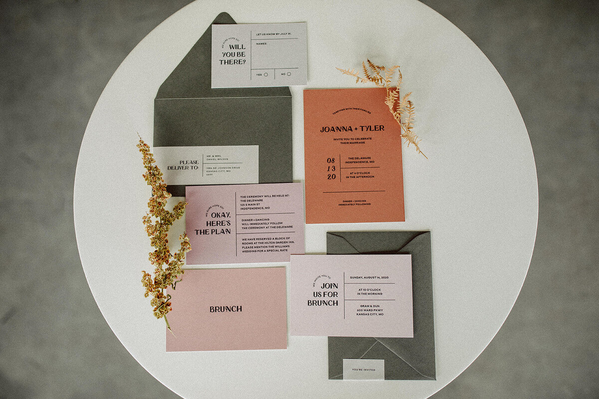 Light gray, peach and mauve-colored wedding stationery with black font atop a white table with plant stems.