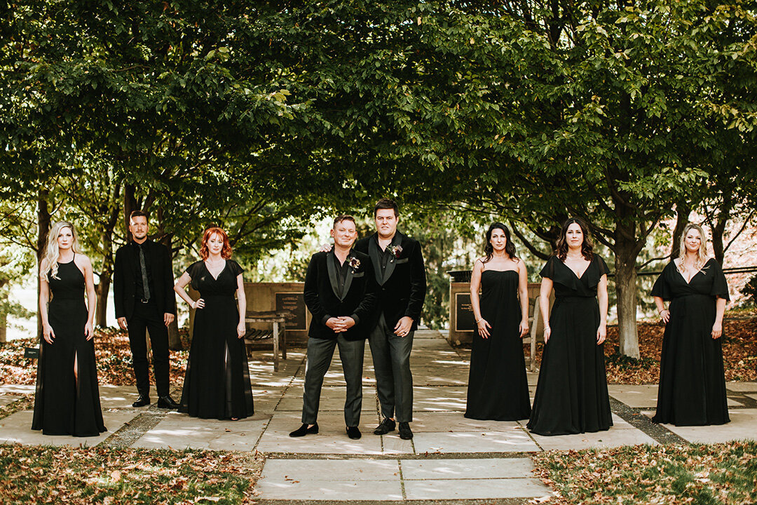 Two grooms wearing black tuxedos pose with their wedding party outdoors.