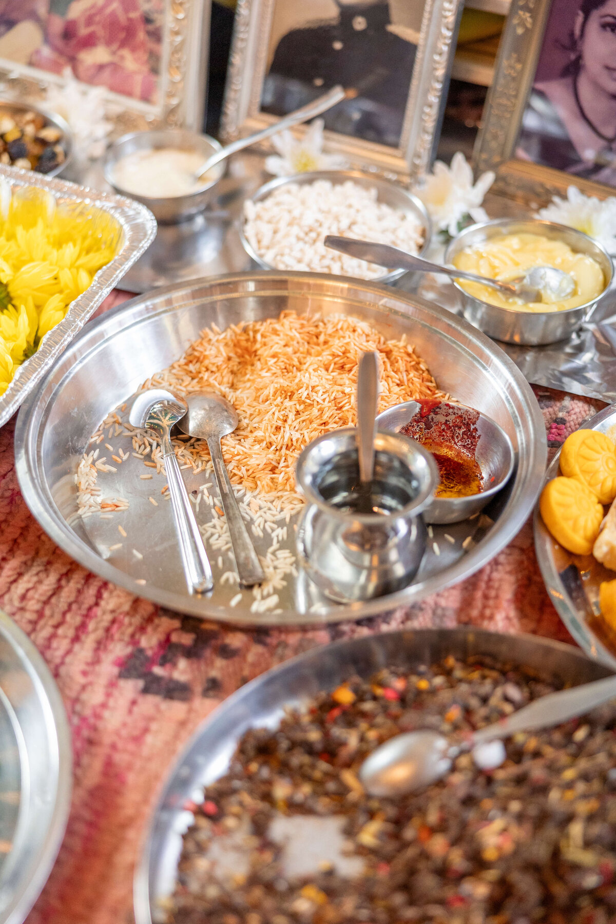 A variety of Indian dishes are presented on a table, including a large metal plate with rice, a sweet dish, and two metal cups. Additional trays hold different foods like lentils and desserts. Decorative frames display faded photos in the background.