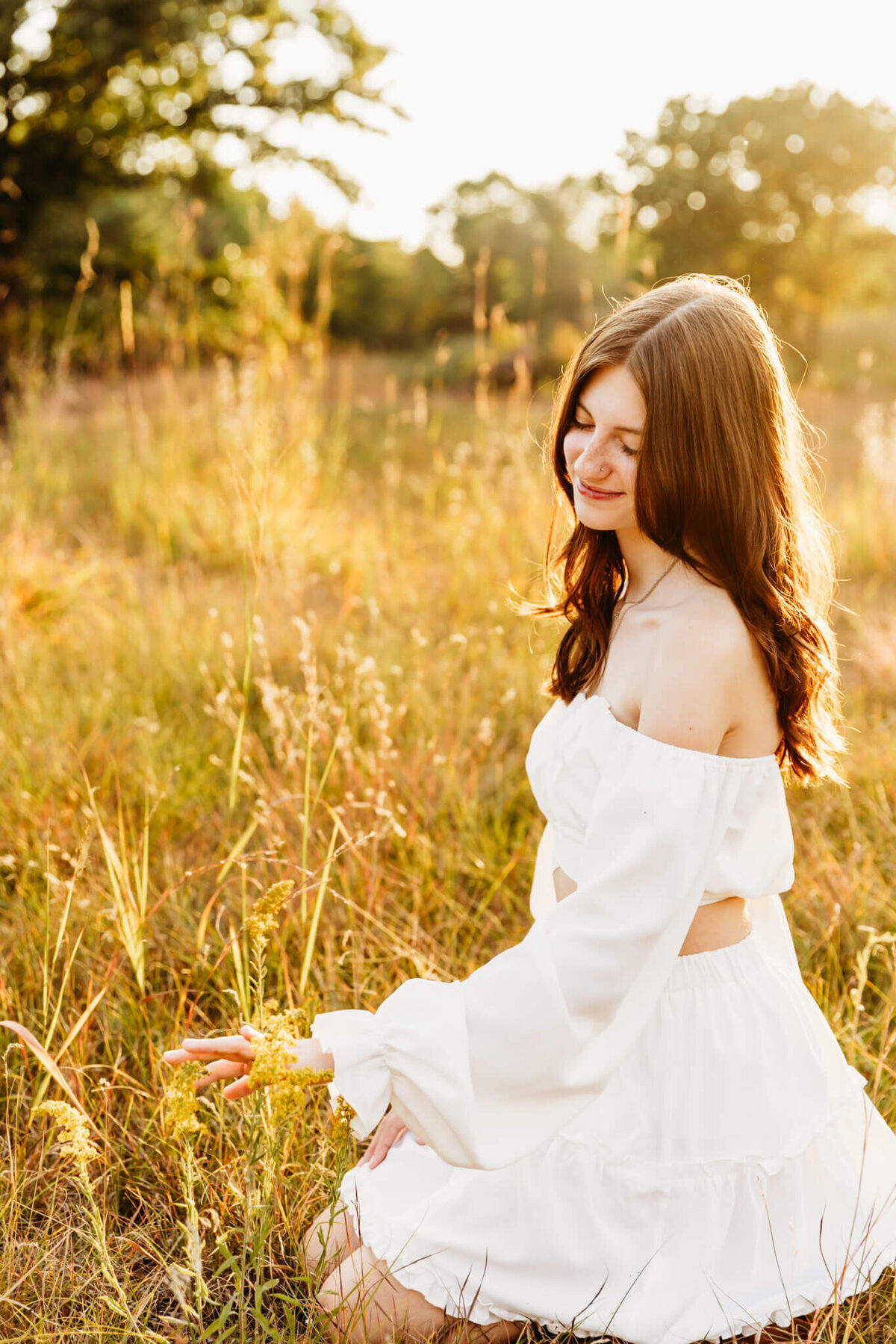 senior girl in a white dress kneeling next to grass at sunset by Ashley Kalbus Photography