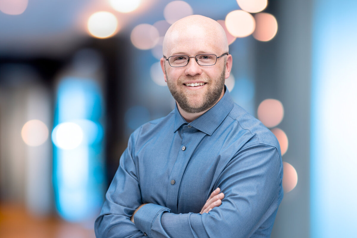 A bald white man real estate professional and realtor with a blue shirt and folded arms poses for a professional headshot portrait at Janel Lee Photography studios in Cincinnati Ohio in an office setting