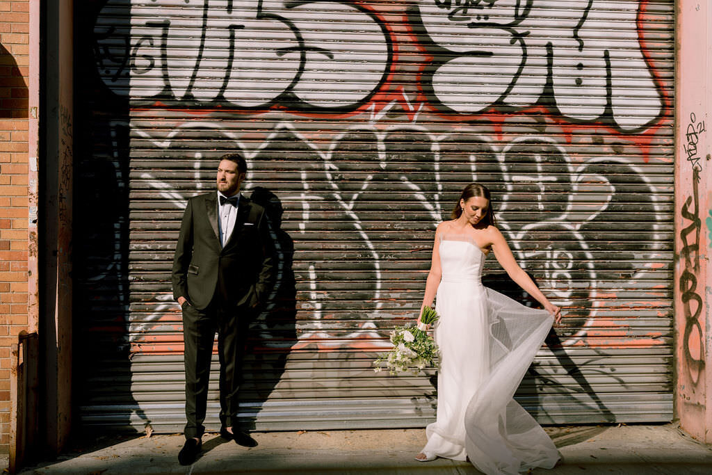 bride and groom standing in front of a graffiti wall while she holds up the side of her dress