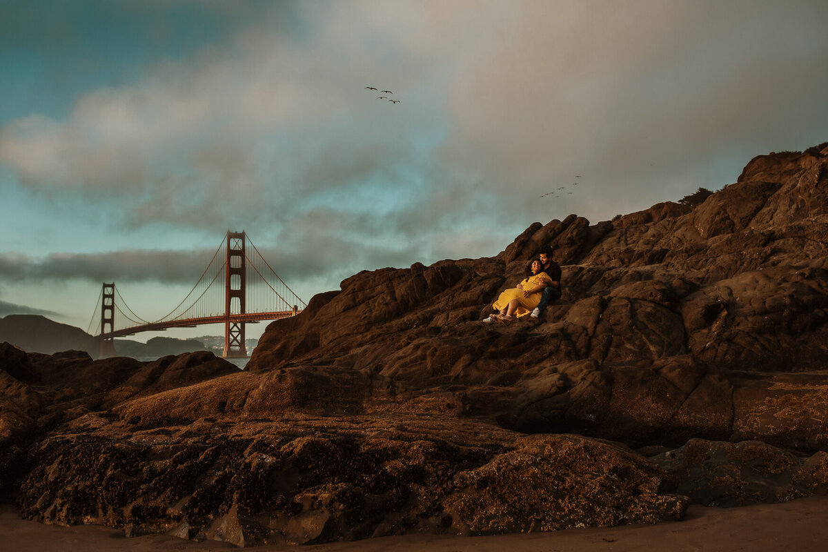 Bay area maternity photography at Baker Beach.  Couple sits atop rocks with Golden Gate Bridge in backdrop.  Pregnant mom in wearing yellow dress