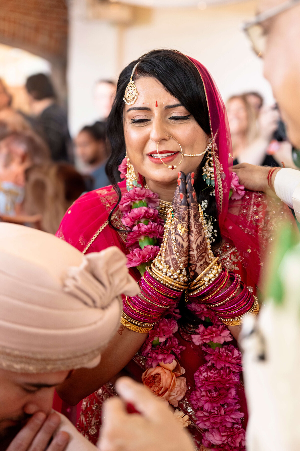 A bride in a red sari with intricate henna and gold jewelry clasps her hands in joy. Shes adorned with garlands, surrounded by well-wishers. The background shows people in festive attire.