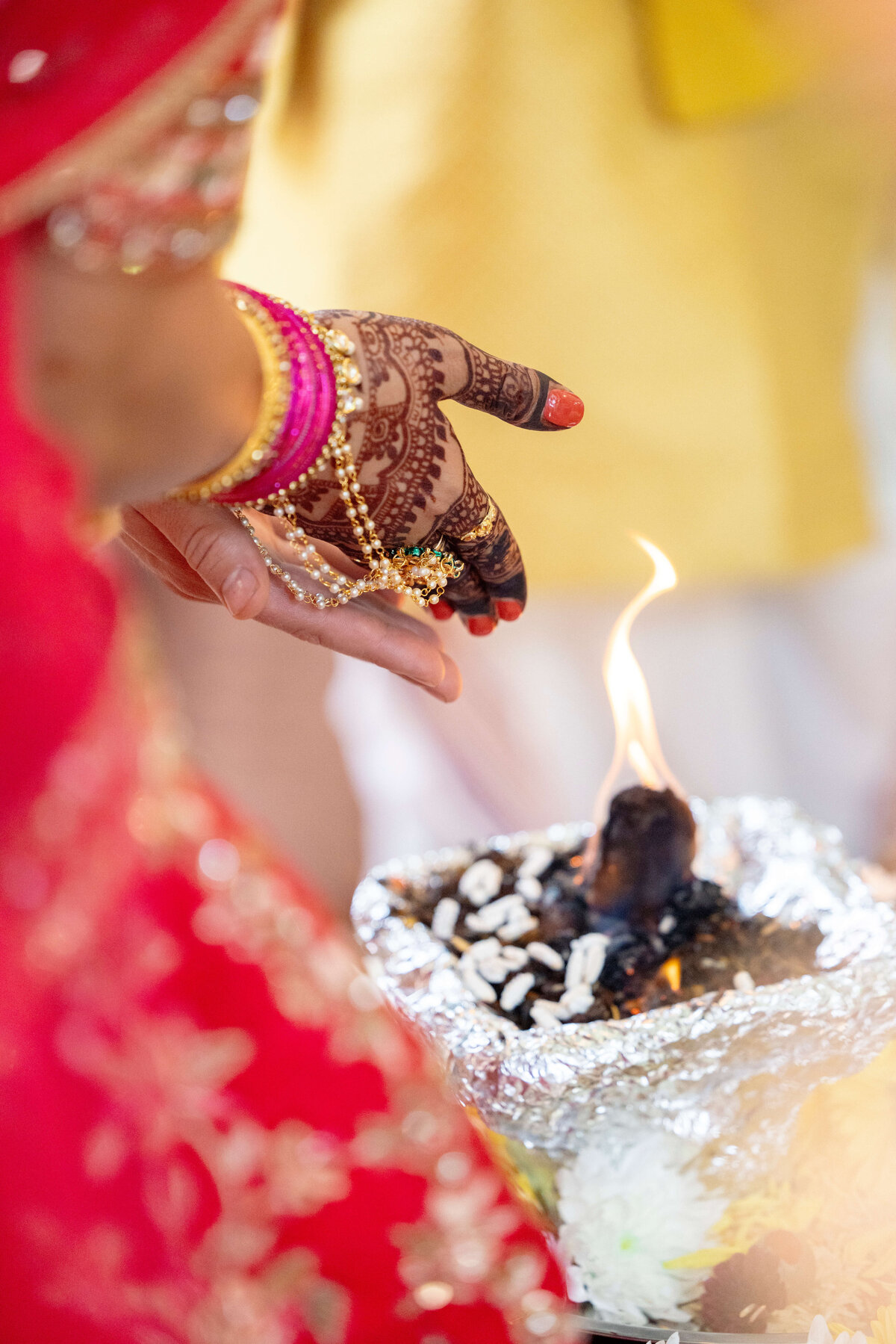 A close-up of a hand adorned with intricate henna designs and gold jewelry, extended over a ceremonial fire. The person is wearing a red garment, and the scene captures part of a Hindu ritual.