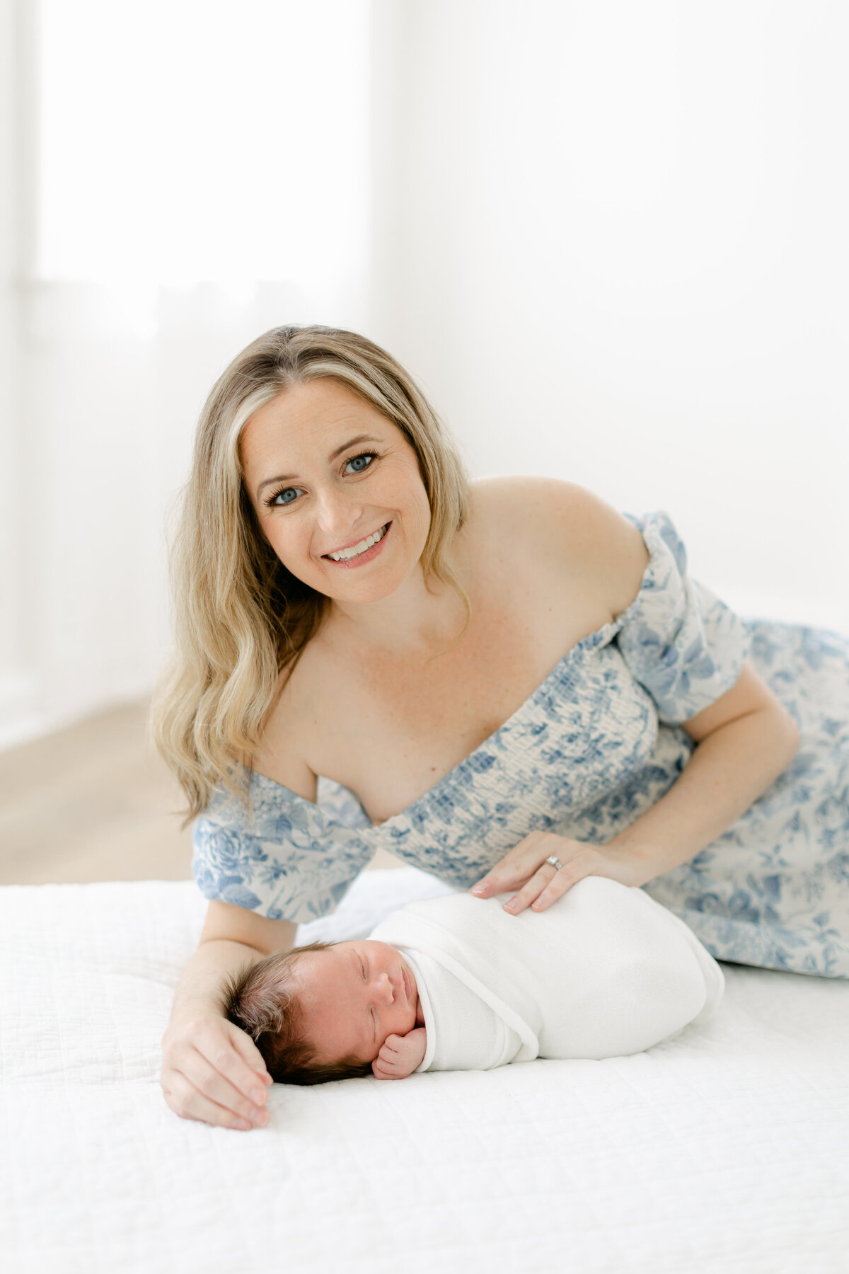 mom laying on a white bed wearing a stunning blue floral pattern dress holding her newborn baby boy photographed by Philadelphia Newborn Photographer Tara Federico