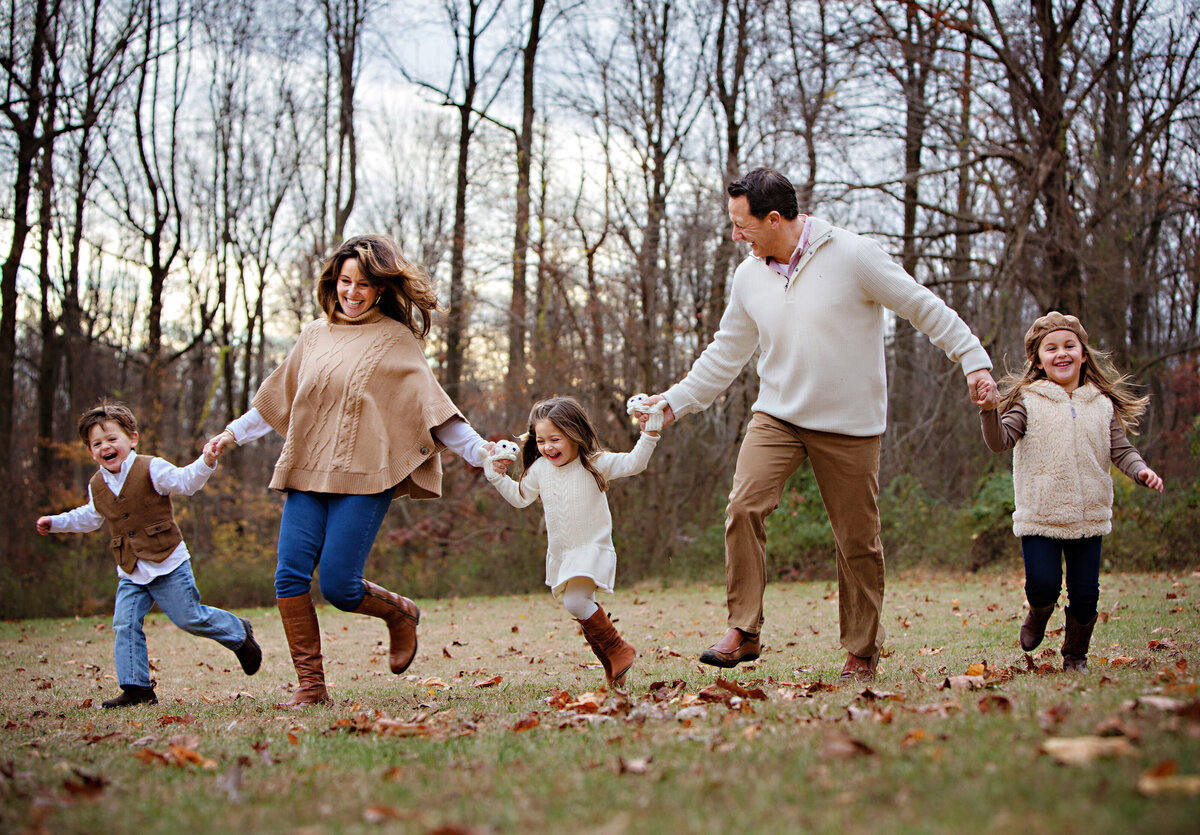 Playful family portrait of parents with young children running through an Autumn field with scattered brown leaves on the ground.  Twin son and daughter along with their older sister are holding hands with Mom and Dad and running together while wearing the perfect fall color scheme of khaki and oatmeal mixed with blue jeans, khaki pants and a sprinkling of tan boots and a tan vest on the son. 