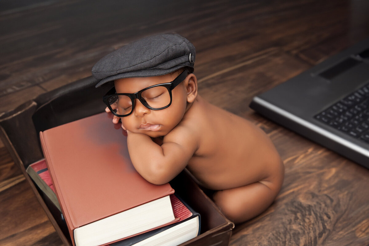 A baby wearing glasses and a flat cap sleeps peacefully against a stack of books, an open laptop nearby. The scene, captured by a Jacksonville newborn photographer, is set against a warm, cozy wooden backdrop.