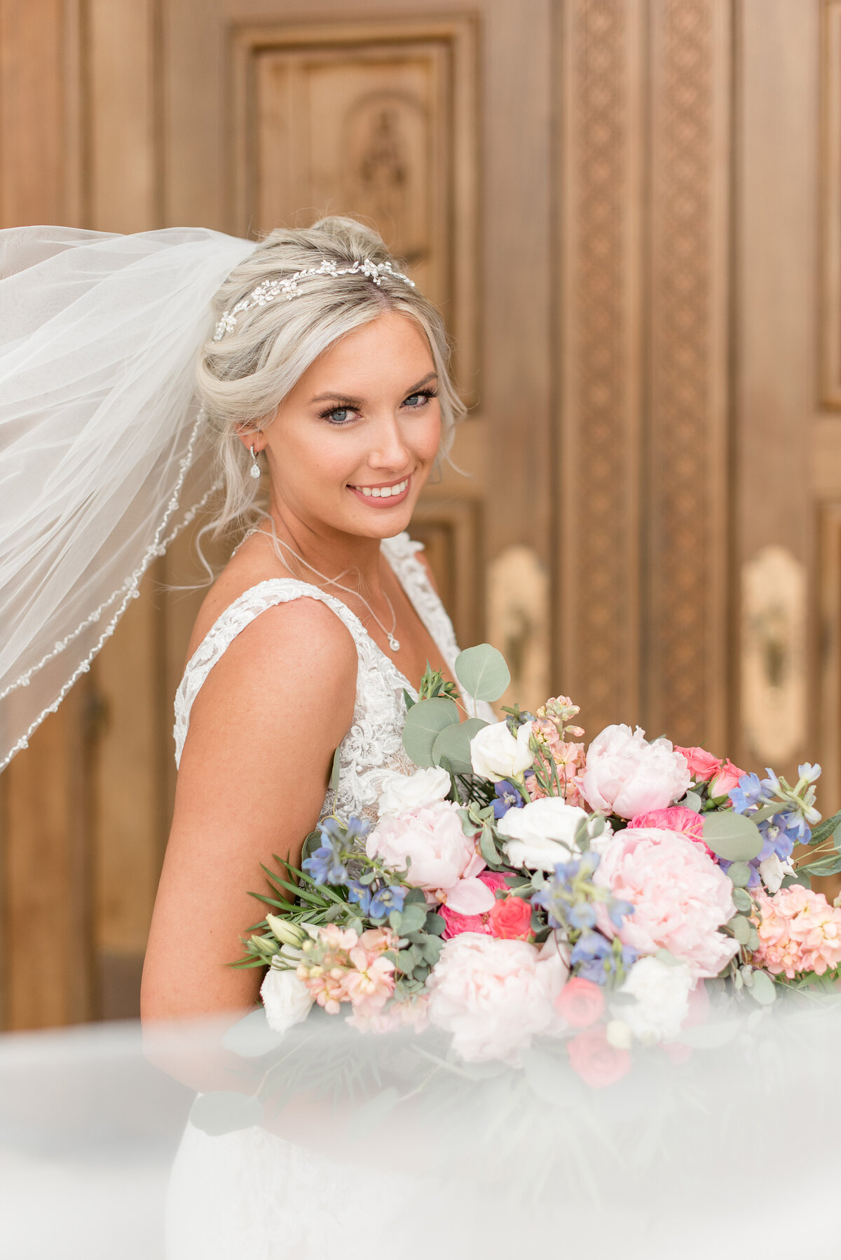 Bride smiling at camera and holding large colorful bouquet filled with peonies, roses, eucalyptus and other flowers.