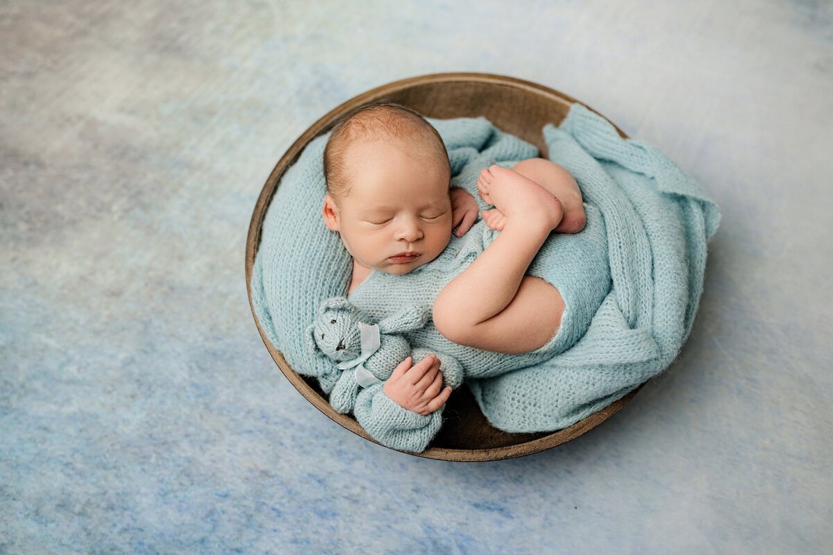 newborn baby boy is dressed in blue posed in a bowl on a blue background