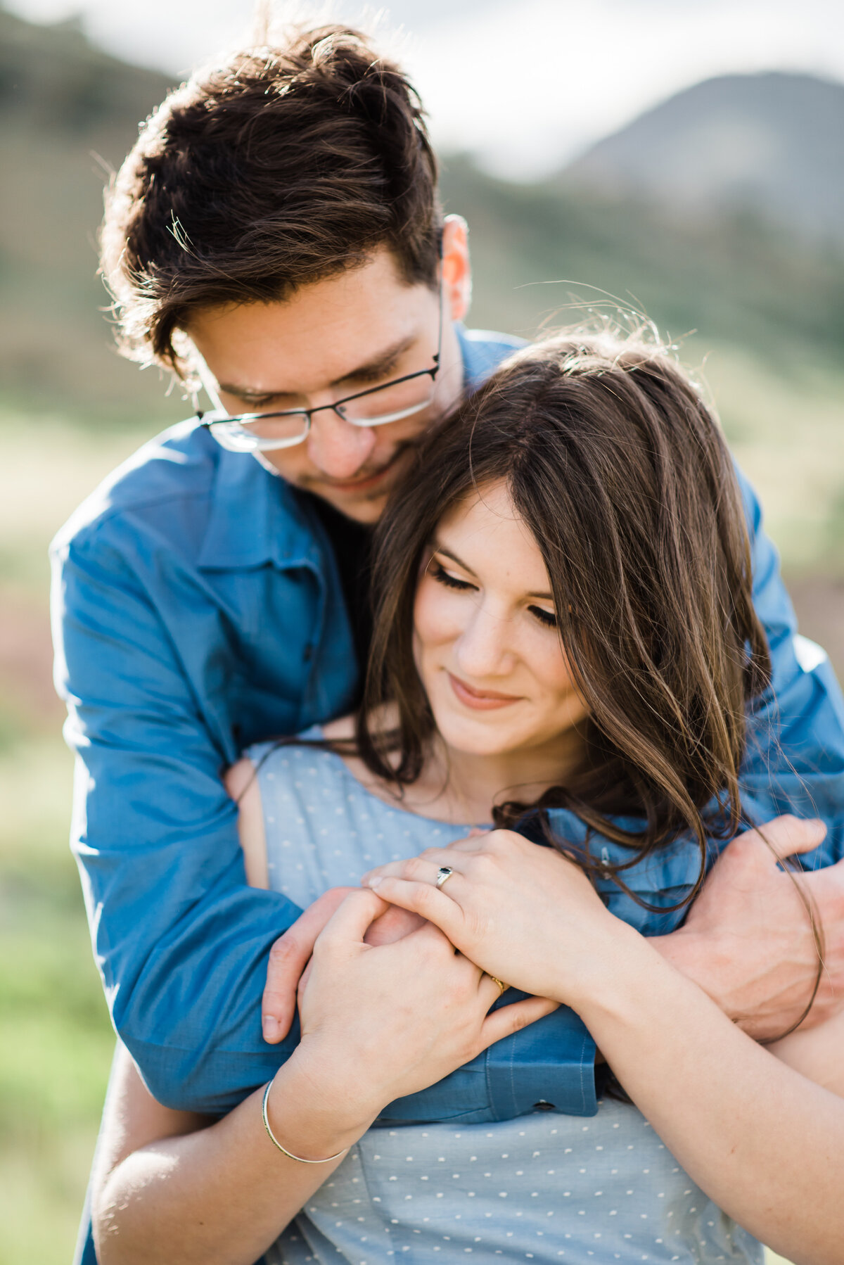 denver engagement photographer captures colorado engagement photos with man standing behind woman while wrapping his arms around the woman's shoulders as she smiles and looks down