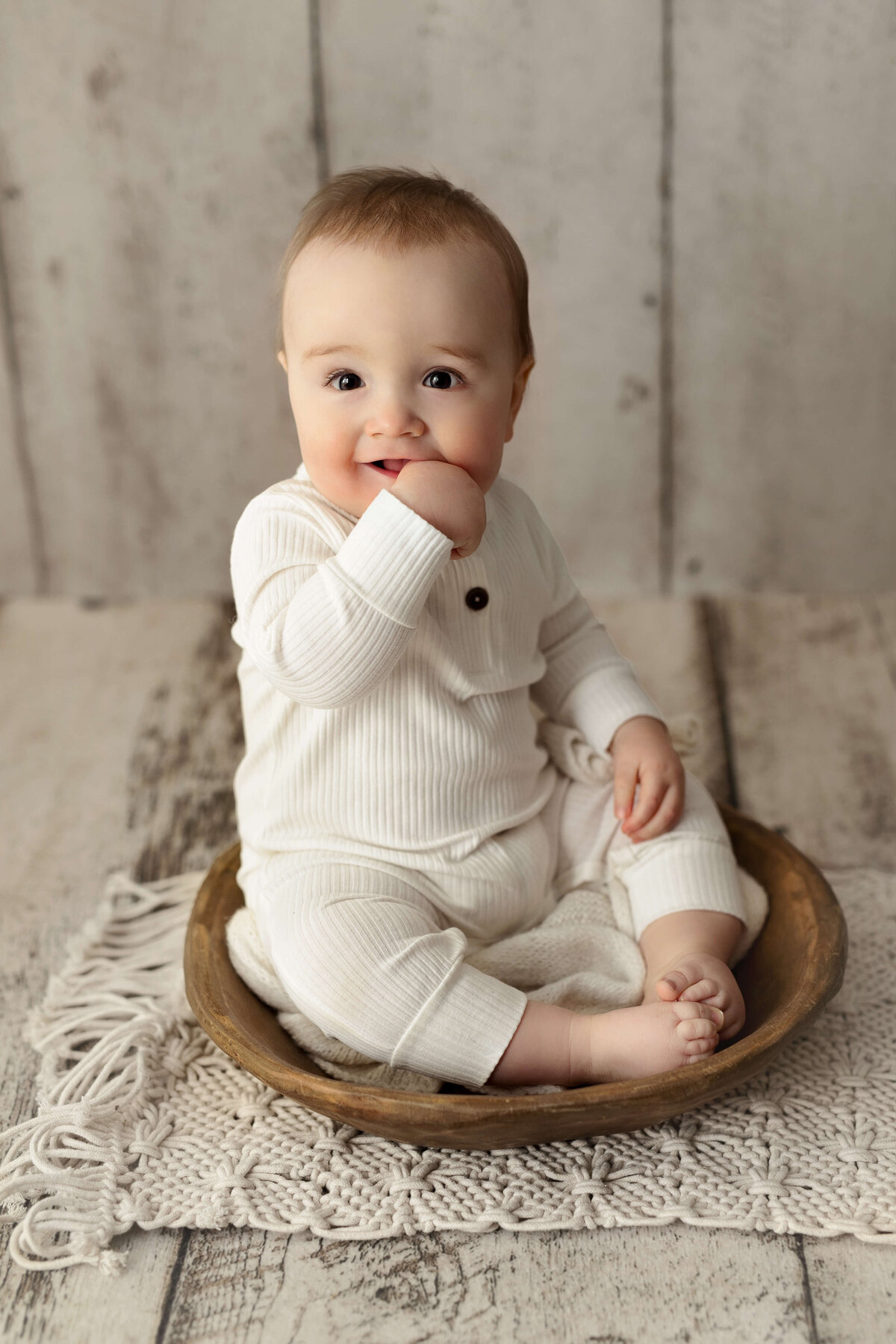 8 month old baby in a white onesie sitting in a wooden bowl on a white wooden background during a toddler milestone session