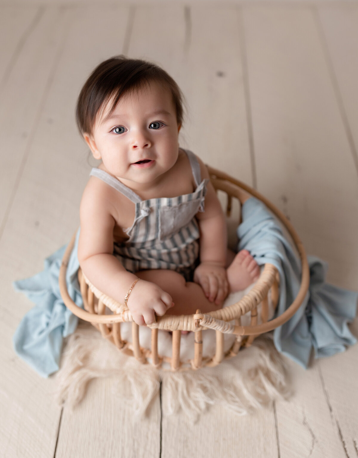 Baby boy sitting in a rattan basket for 6-month milestone photography session at West Palm Beach photography studio. Baby is looking up at the camera with dark brown hair. Baby is wearing striped short overalls. Basket is on painted barn board floors.