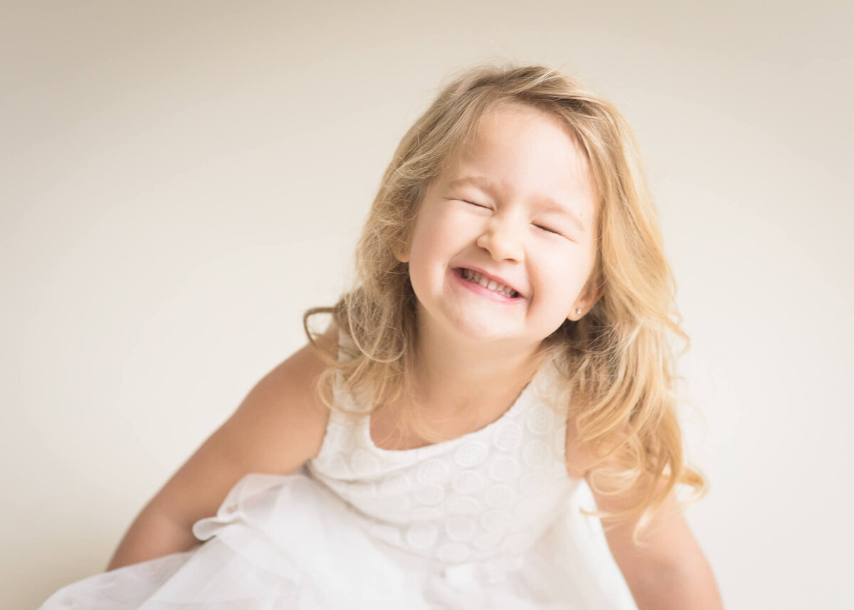 children's studio portrait of young girl in white being so silly showing persoality with a light backdrop behind her