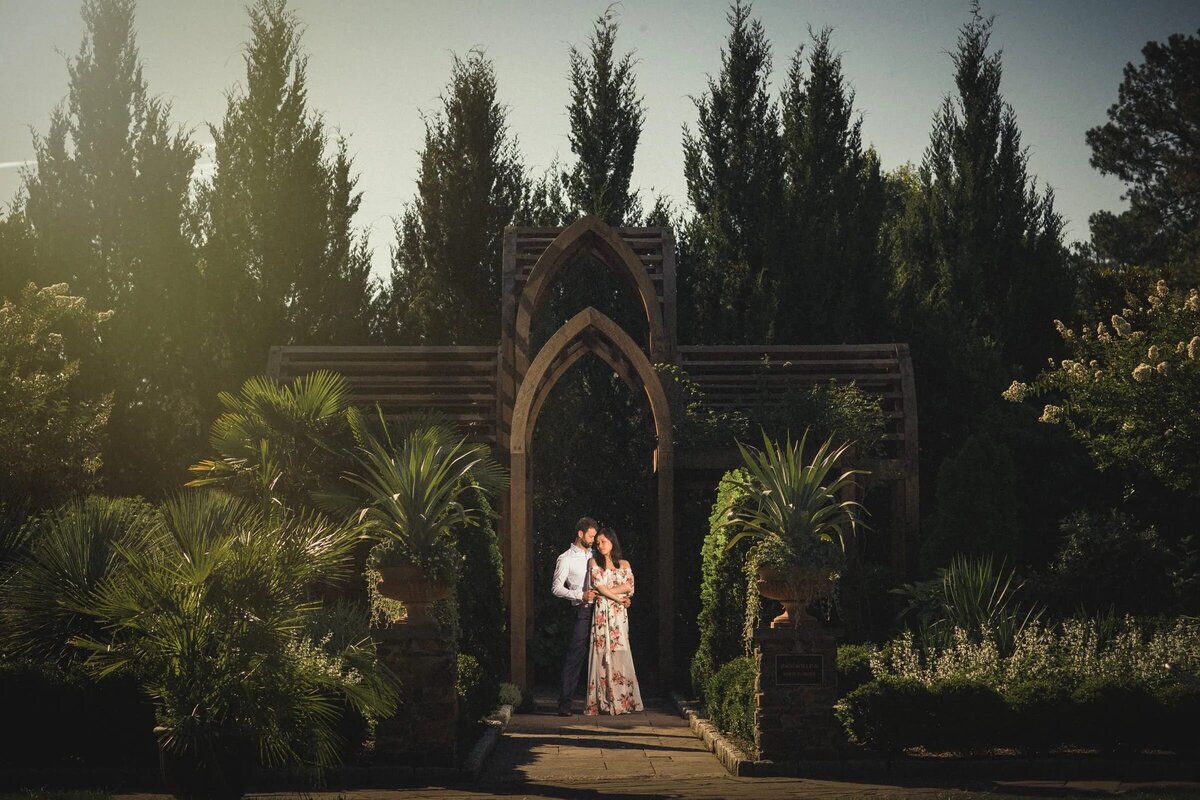 Couple stands in a gothic archway within a lush garden, the sun casting a warm glow