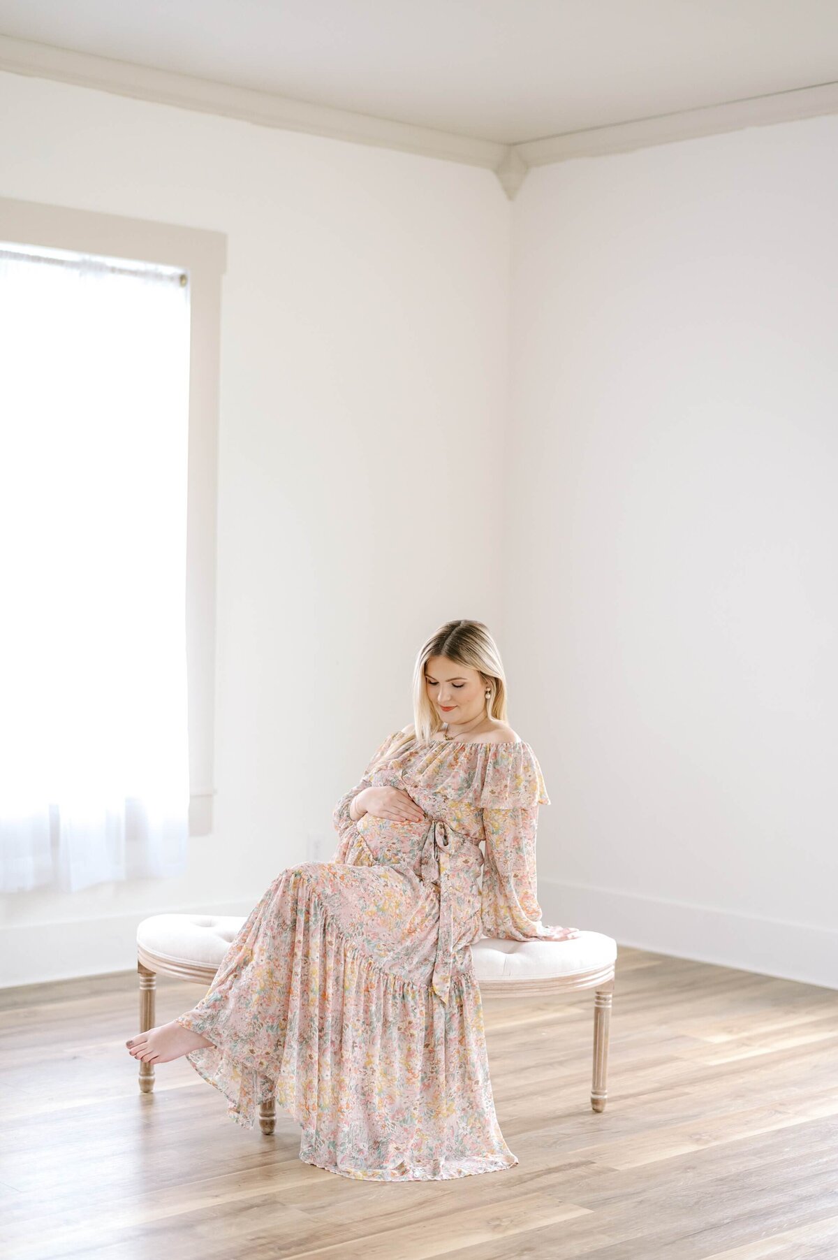 An expecting mother sits on a bench with her legs crossed in a Charlotte, NC studio captured by Charlotte Maternity Photographer Melissa Mayrie Photography.