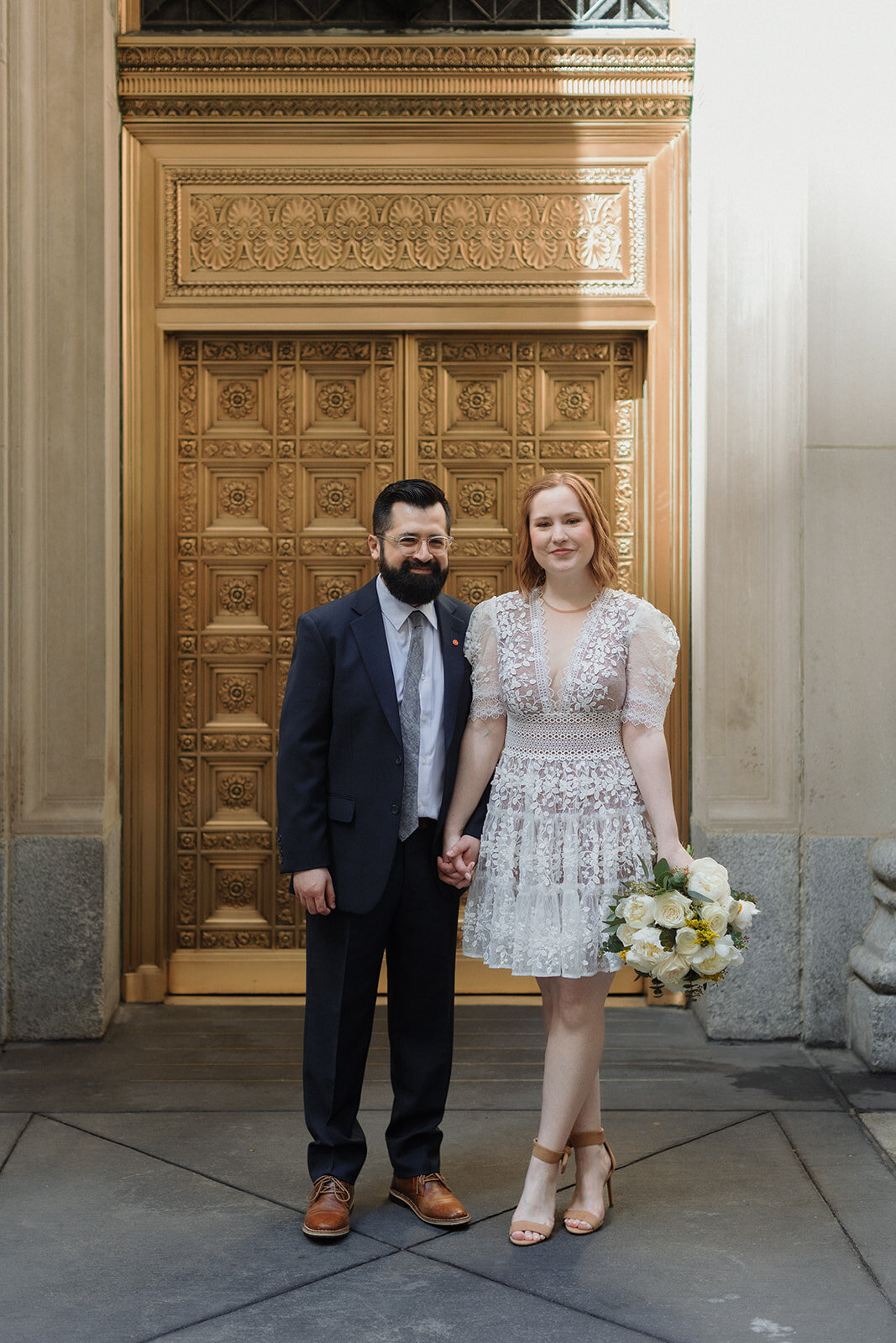 Wedding couple holds hands and stands facing camera in front of large gold brass doors are behind them.