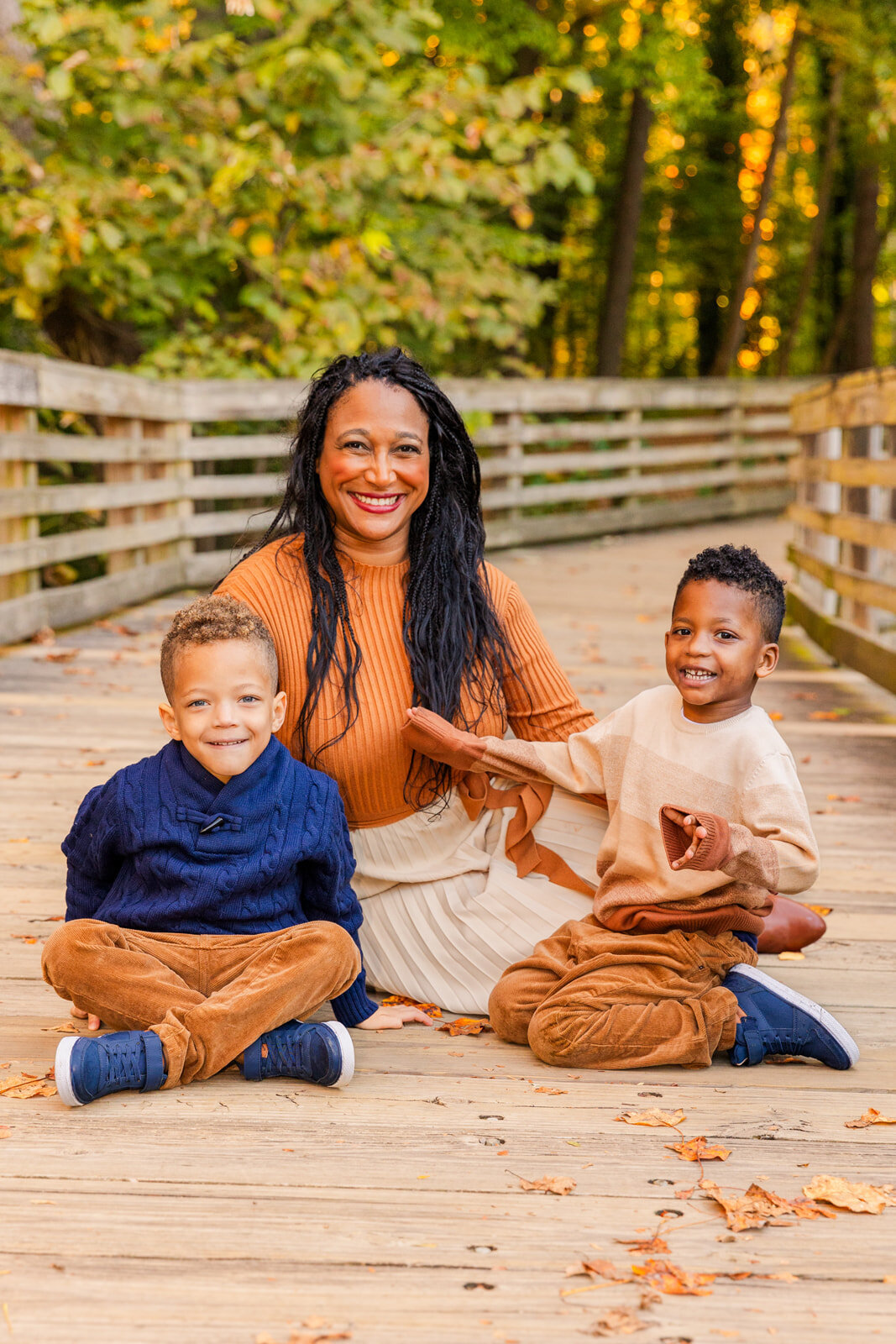 Mom sitting on a bridge with her 2 toddler sons wearing coordinated orange and blue outfits during a Fall photoshoot in Atlanta with Laure Photography