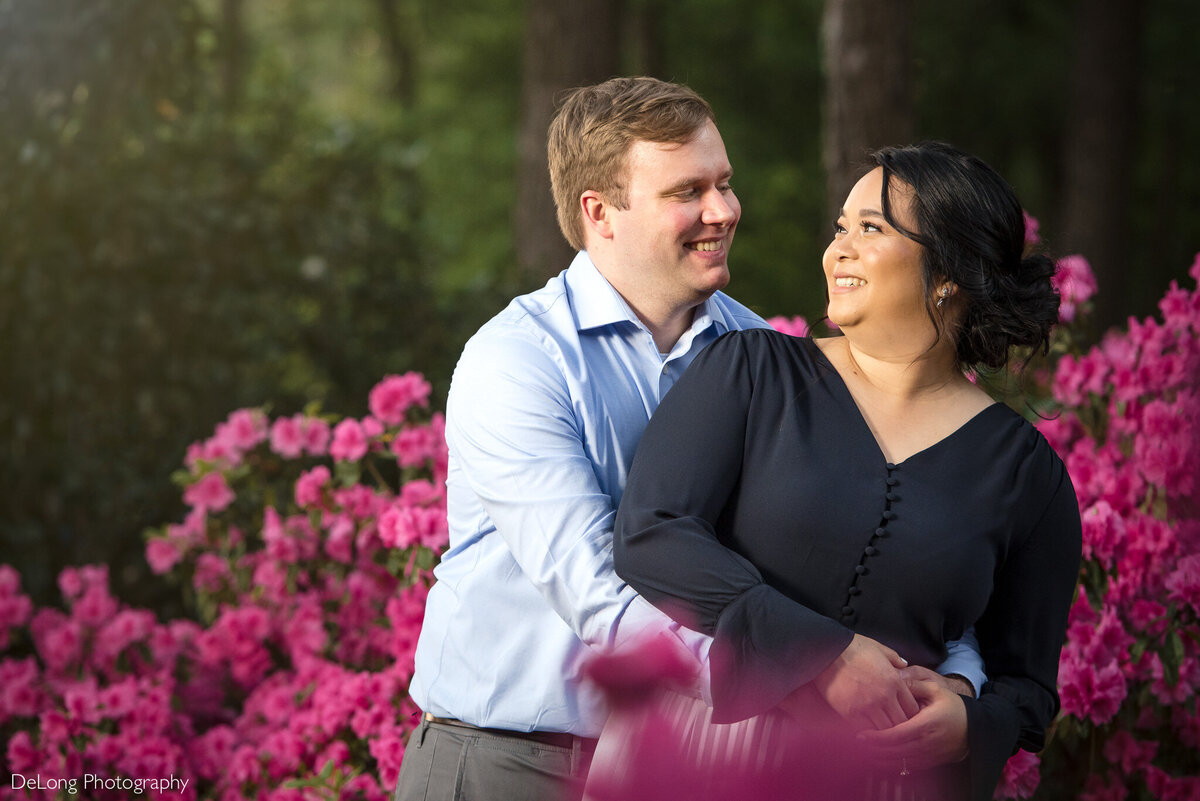 Man-standing-behind-his-fiancee-with-his-arms-wrapped-around-her-smiling-at-each-other-amid-pink-spring-flowers-at-Jetton-Park