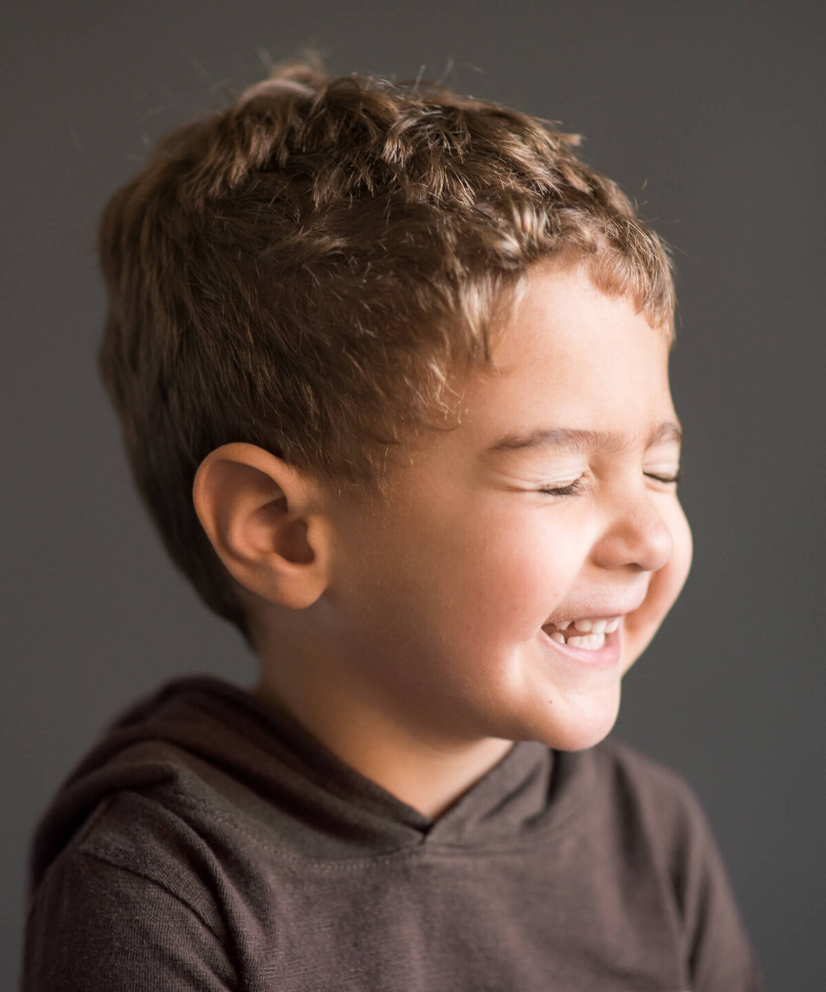 super cute little boy  profile of his face while laughing with eyes closed, grey backdrop studio portrait