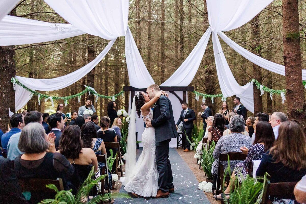 Bride and groom, sharing a first kiss in a forest while their wedding guests cheer and clap