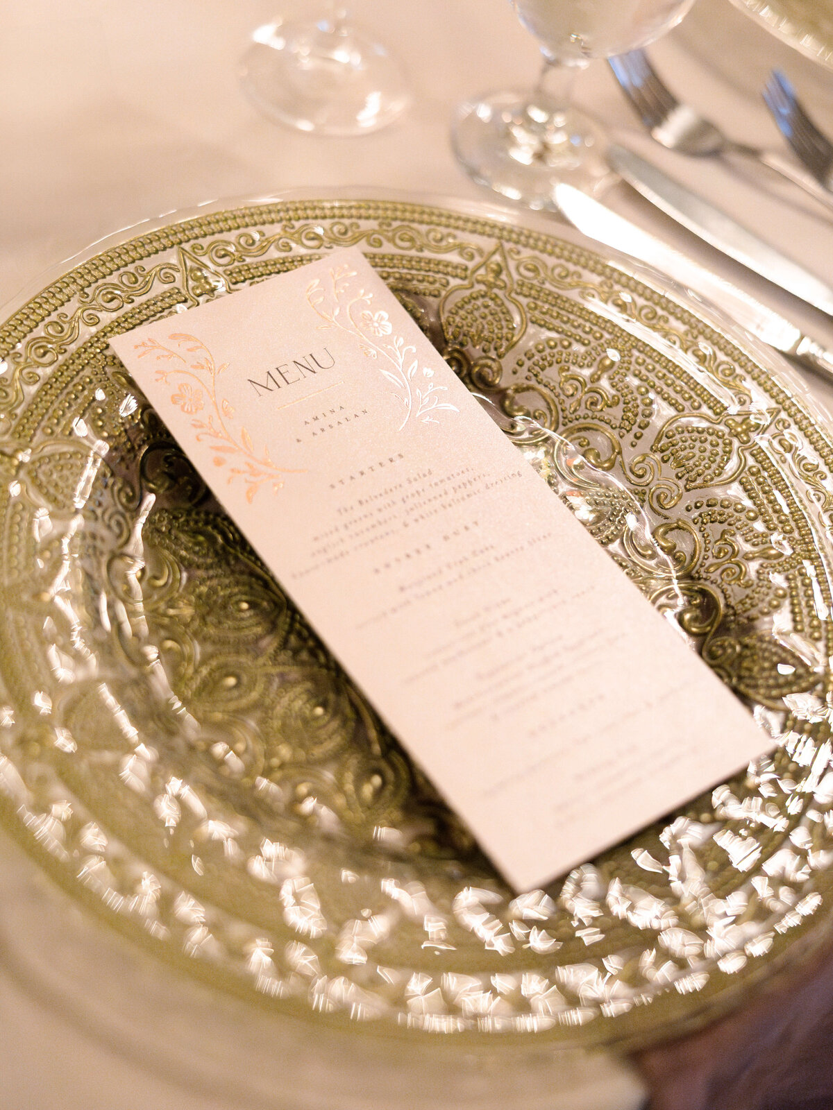 A decorative table setting featuring an ornate gold charger plate with intricate patterns, topped with a folded menu card. The menu has elegant, embossed floral designs at the top. A knife and glass are placed beside the plate on a white tablecloth.